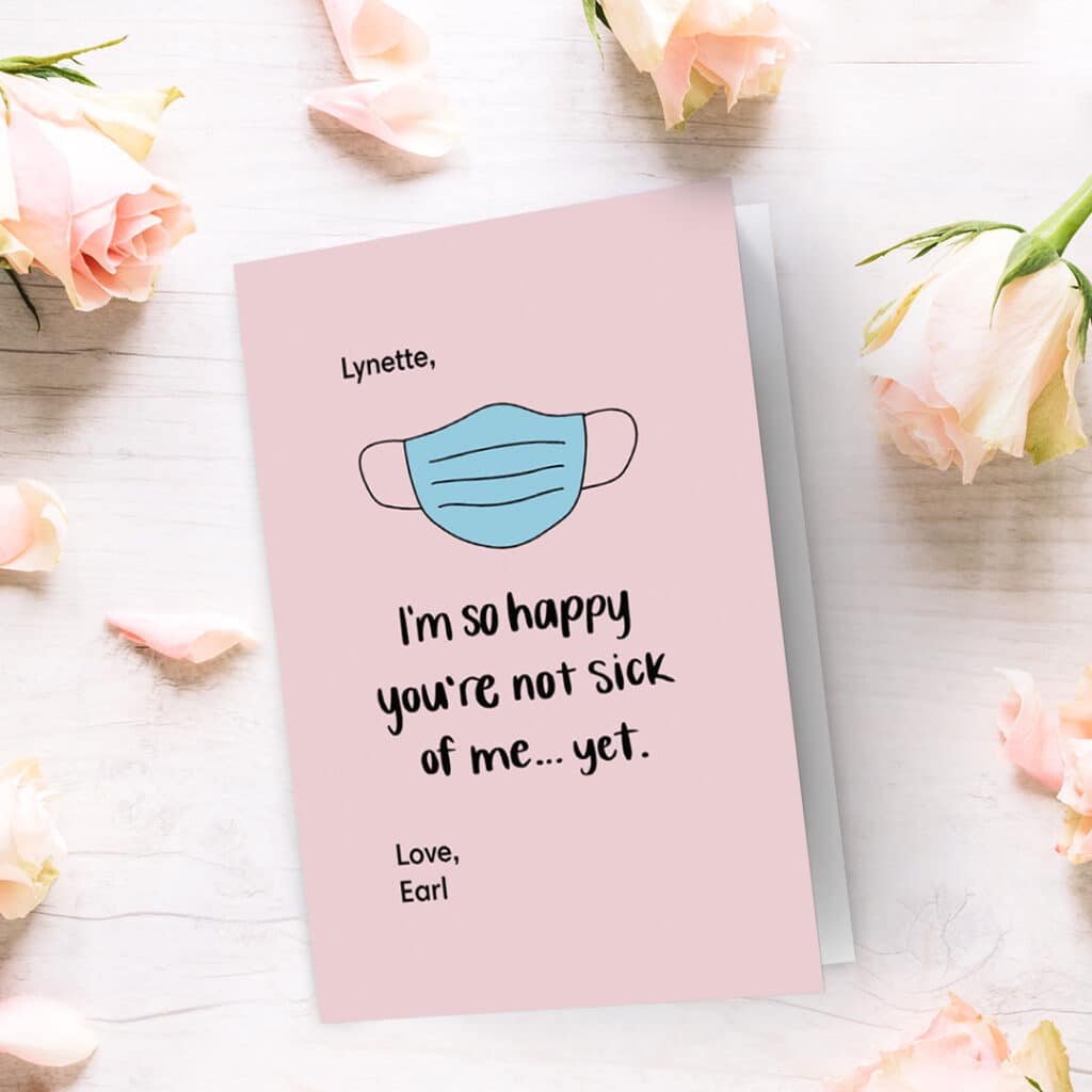 A fun, pink design of a Valentine's day card presented on a surface with lovely light pink roses. COVID related.